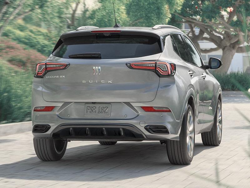 Vue arrière du Buick Encore GX 2025 en mouvement seul sur une route bordée d’arbres.