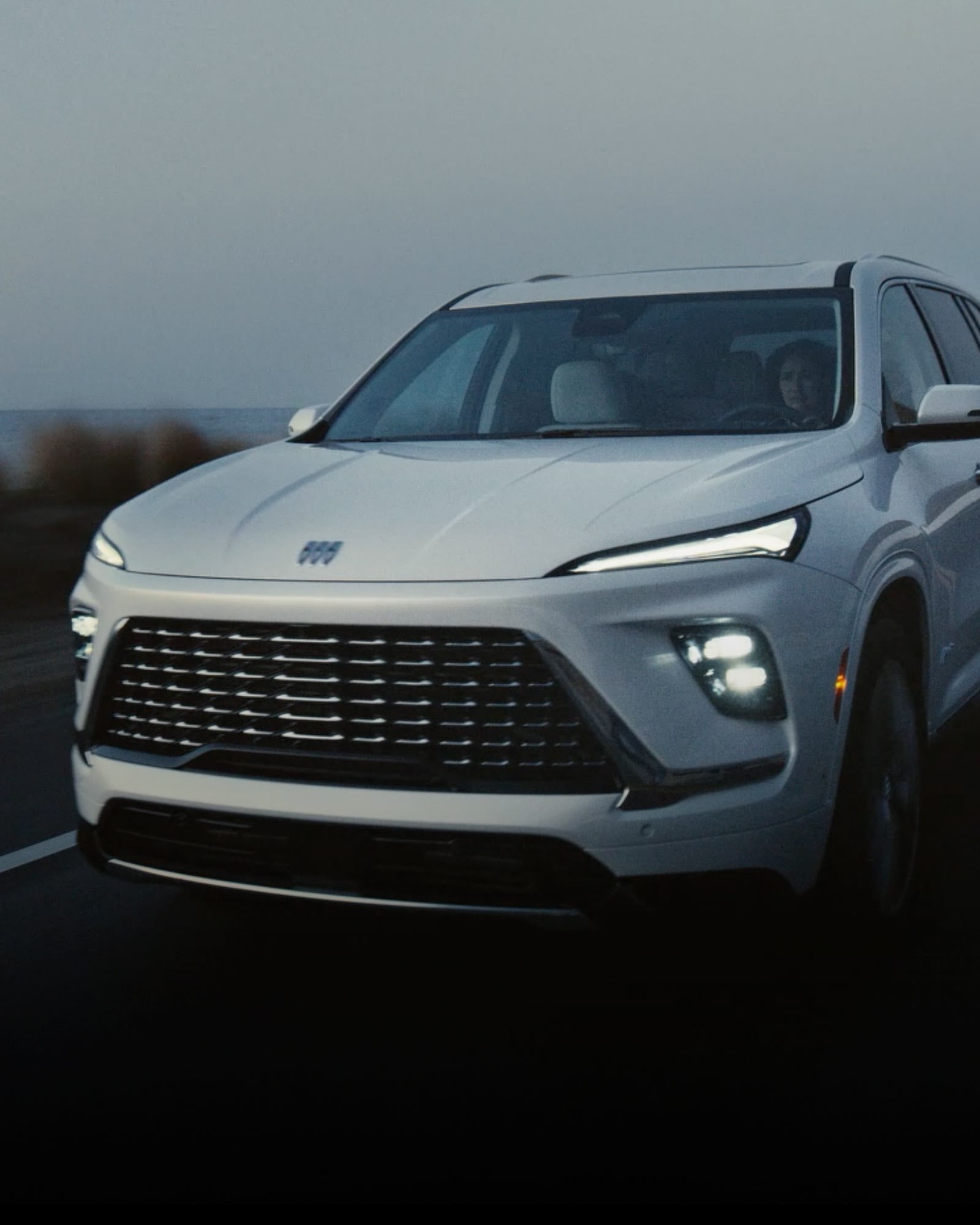 Front view of the 2026 Buick Enclave Avenir driving on the road with its headlights on.