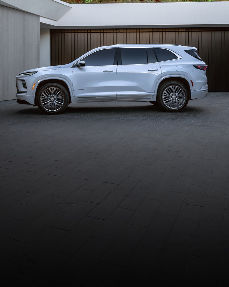 Vue latérale d’un VUS blanc Buick Enclave Avenir 2027 stationné à côté d’un bâtiment moderne aux portes de garage sombres.