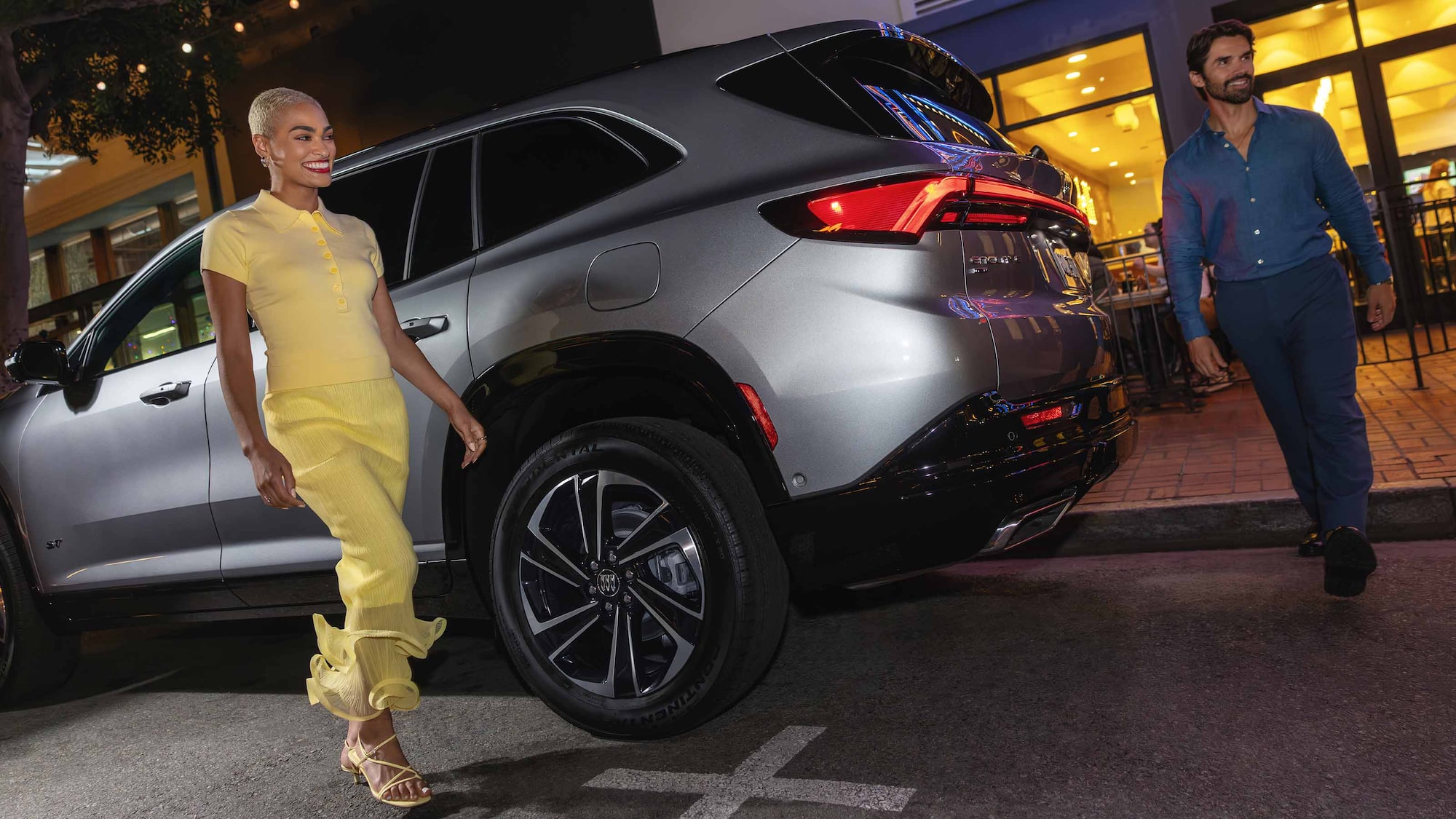 Un homme et une femme passant devant un VUS Buick Enclave gris 2027 dans une rue de la ville la nuit