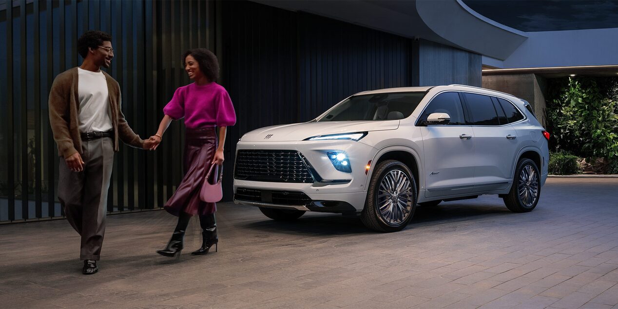 Deux personnes passant devant une Buick Enclave 2027 garées devant une maison moderne la nuit.