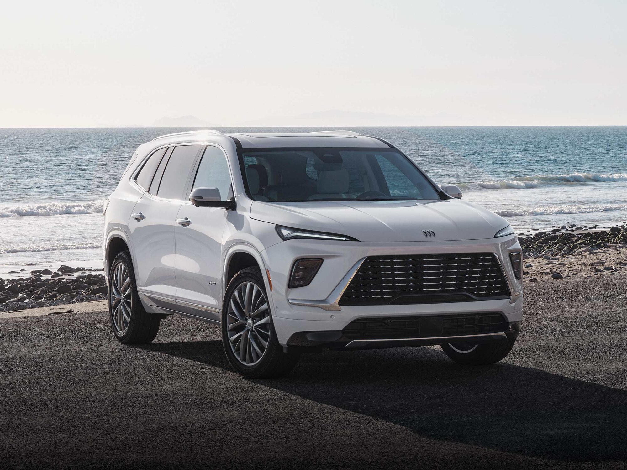 Front Three-quarters View of a White Buick Enclave Parked on a Beach as Waves Crash on the Shoreline