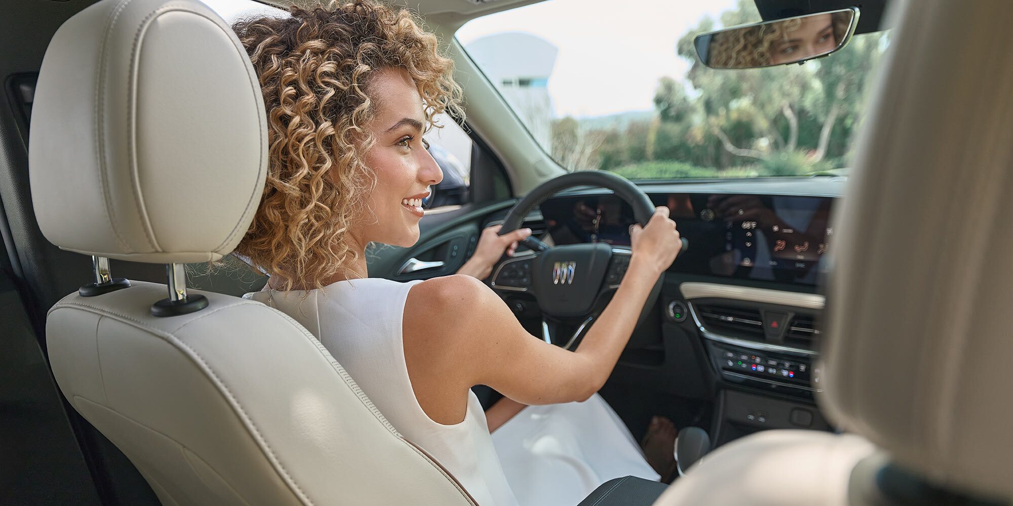 A Smiling Woman Drives the Buick Encore GX in a White Dress with Her Hands on the Steering Wheel