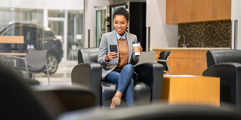 A Customer Viewing Her Phone while Holding a Coffee and Sitting in a Sofa at a Buick Dealership