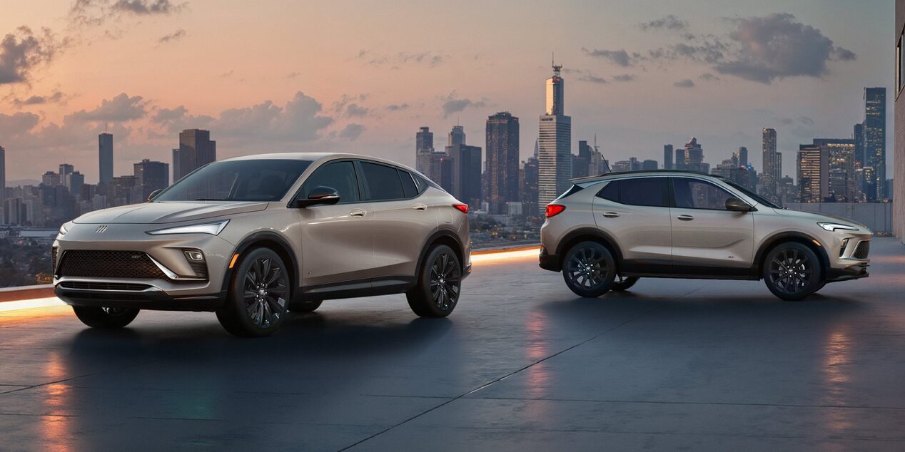 The 2026 Envista Sport Touring SUV and the 2026 Encore GX Sports Touring SUV both in the Color Ceramic Parked on a Parking Garage Rooftop Overlooking a City Skyline