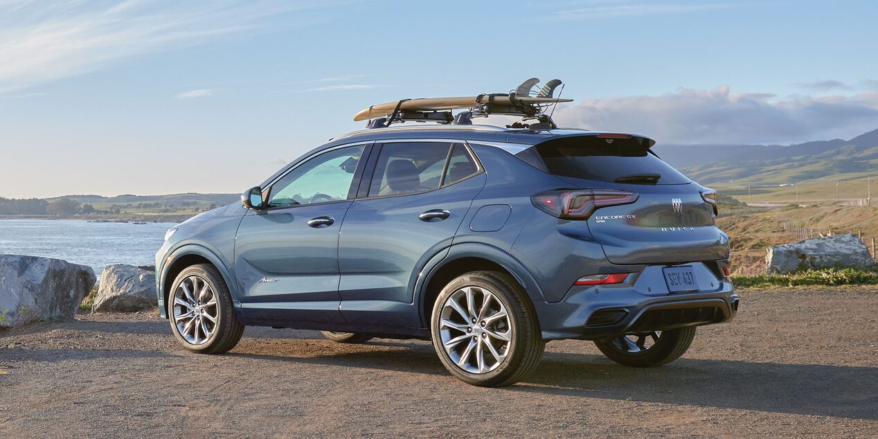 Rear Three-Quarters View of a Buick Encore GX Avenir Luxury SUV Parked Overlooking a Large Body of Water with a Surfboard Attached to the Top of the Vehicle
