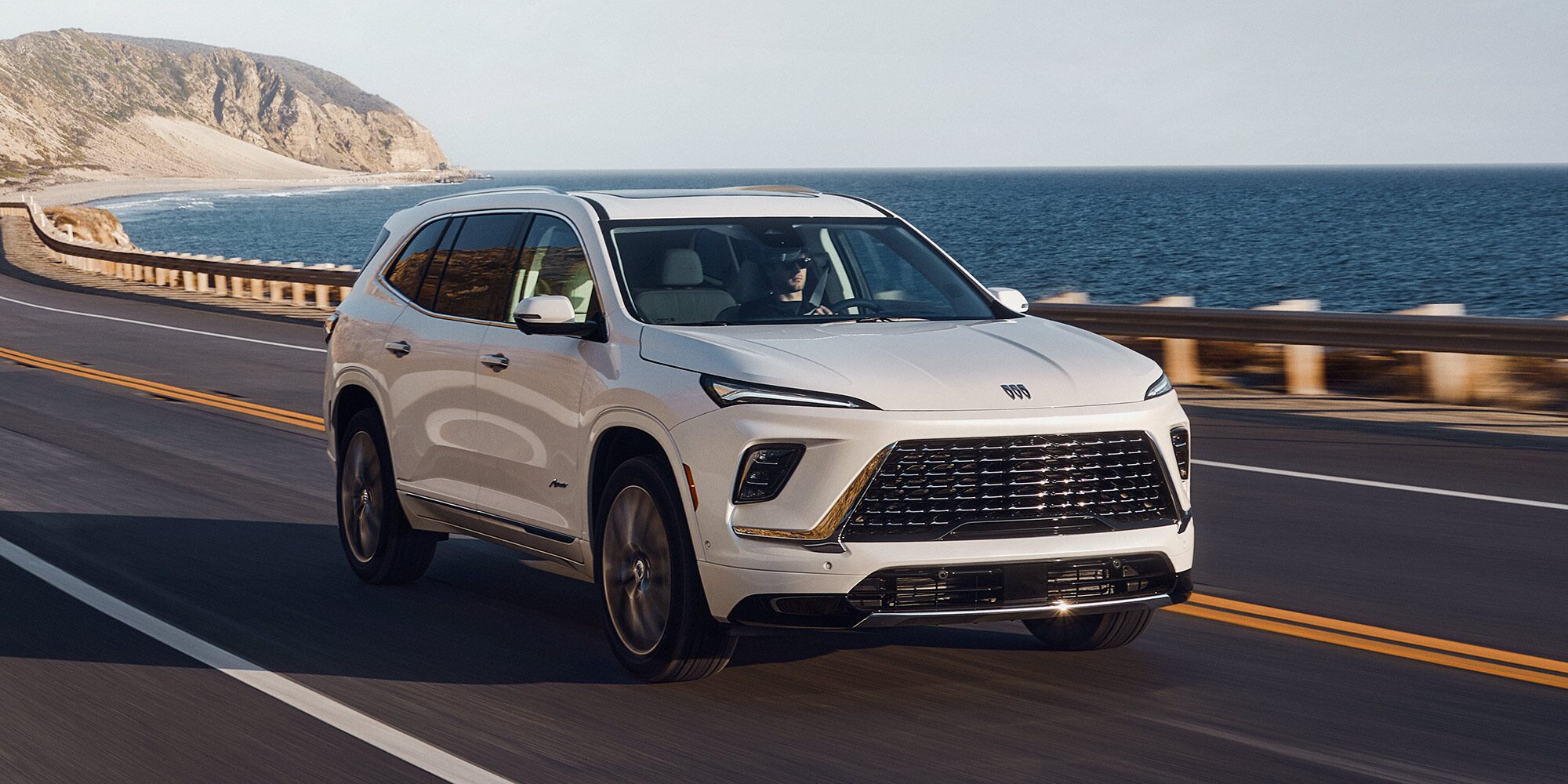 A White Buick Enclave SUV Driving on a Coastal Road Overlooking the Ocean with a Distant Rocky Cliff Under a Clear Sky