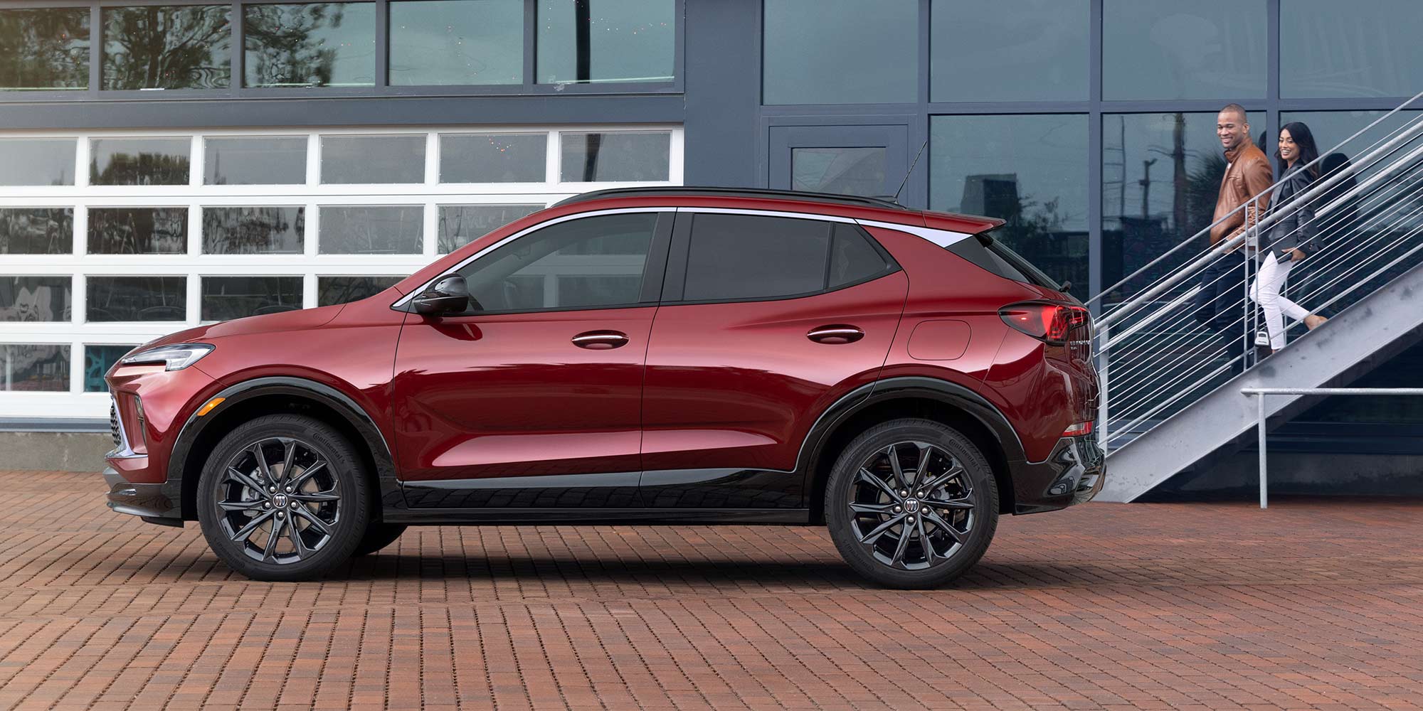 A Dark Red Buick Envista Parked on a Patterned Brick Surface with a Man and Woman Walking Up Stairs in the Background 