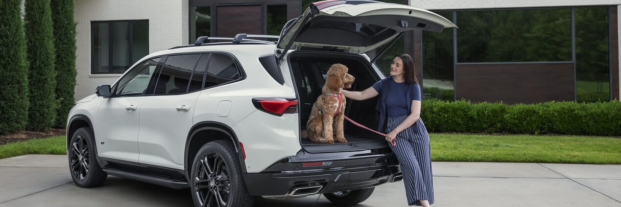 A Woman Sitting Beside the Open Liftgate of a White Buick Enclave SUV Petting a Brown Poodle Wearing a Red Harness