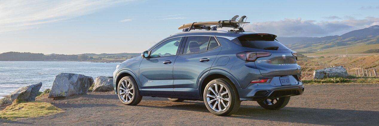 Rear Three-Quarters View of a Buick Encore GX Avenir Luxury SUV Parked Overlooking a Large Body of Water with a Surfboard Attached to the Top of the Vehicle