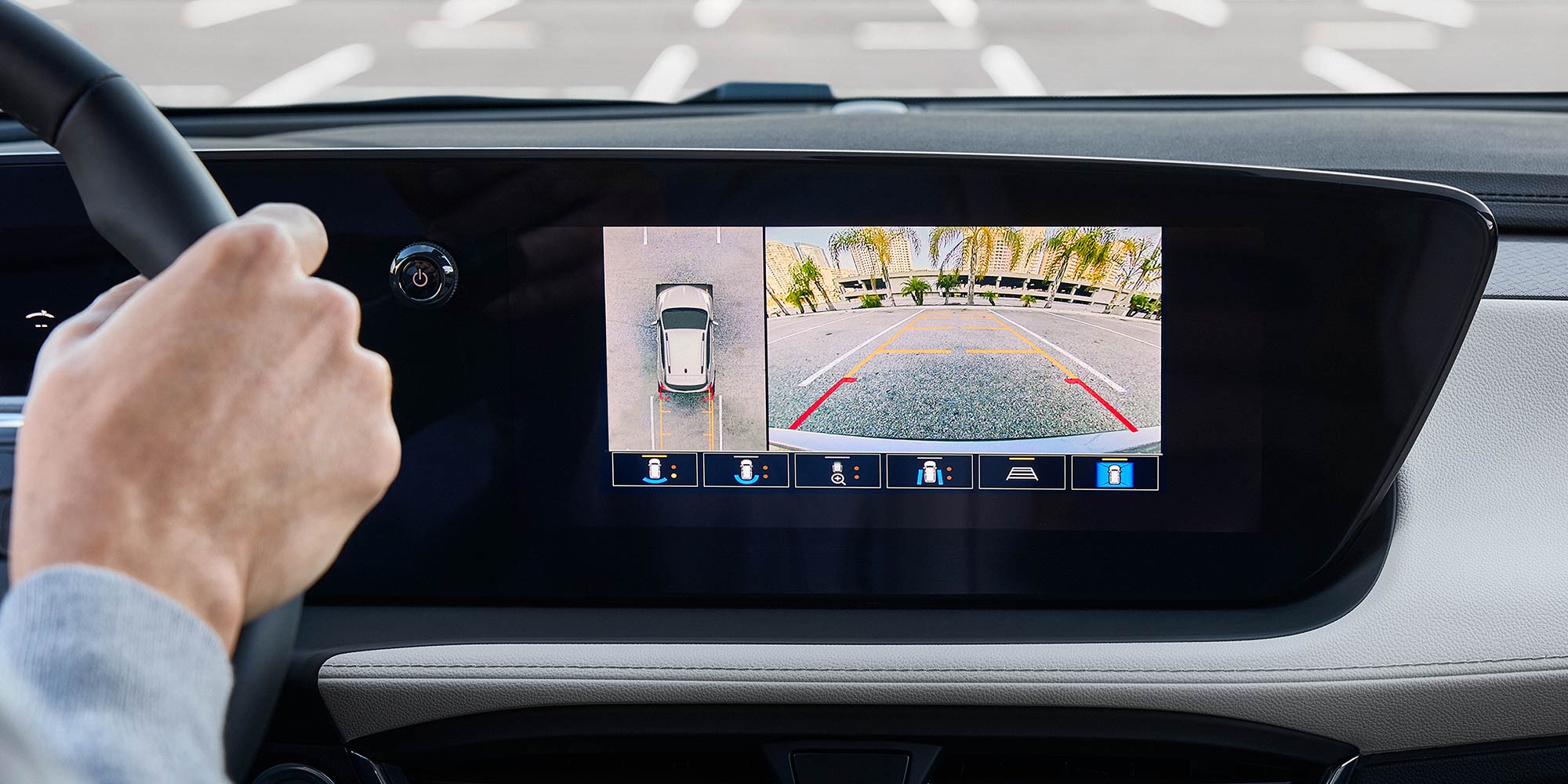A Driver's Hand On the Steering Wheel of a Buick Encore GX with a Camera View On the Dashboard Display of a Parking Lot