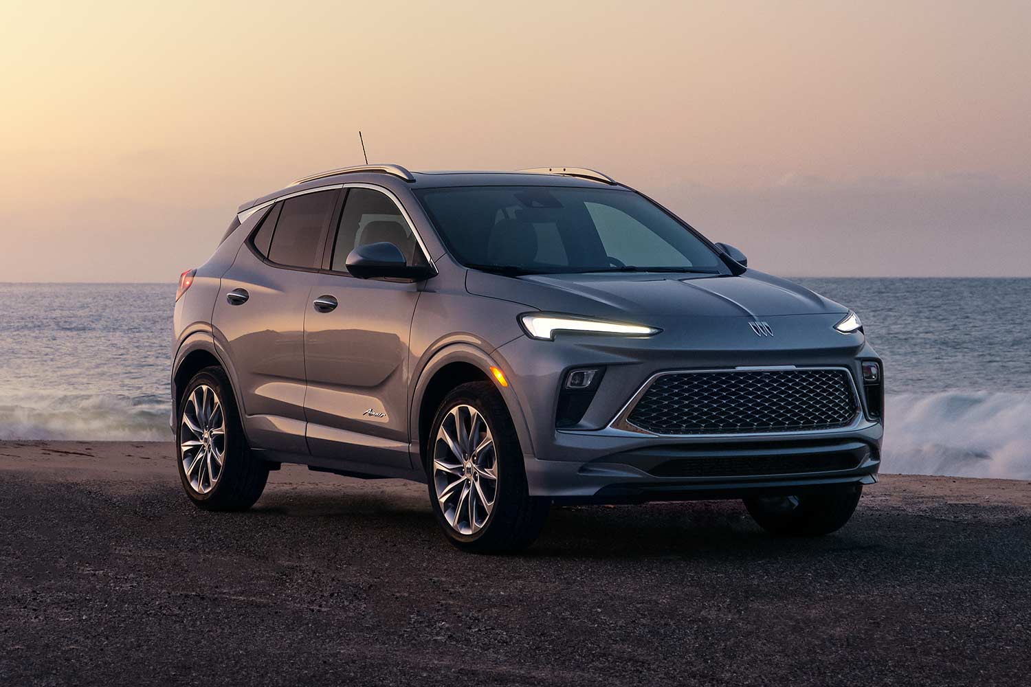 A Gray Buick Encore GX Avenir SUV Parked on a Beach at Sunset with Waves