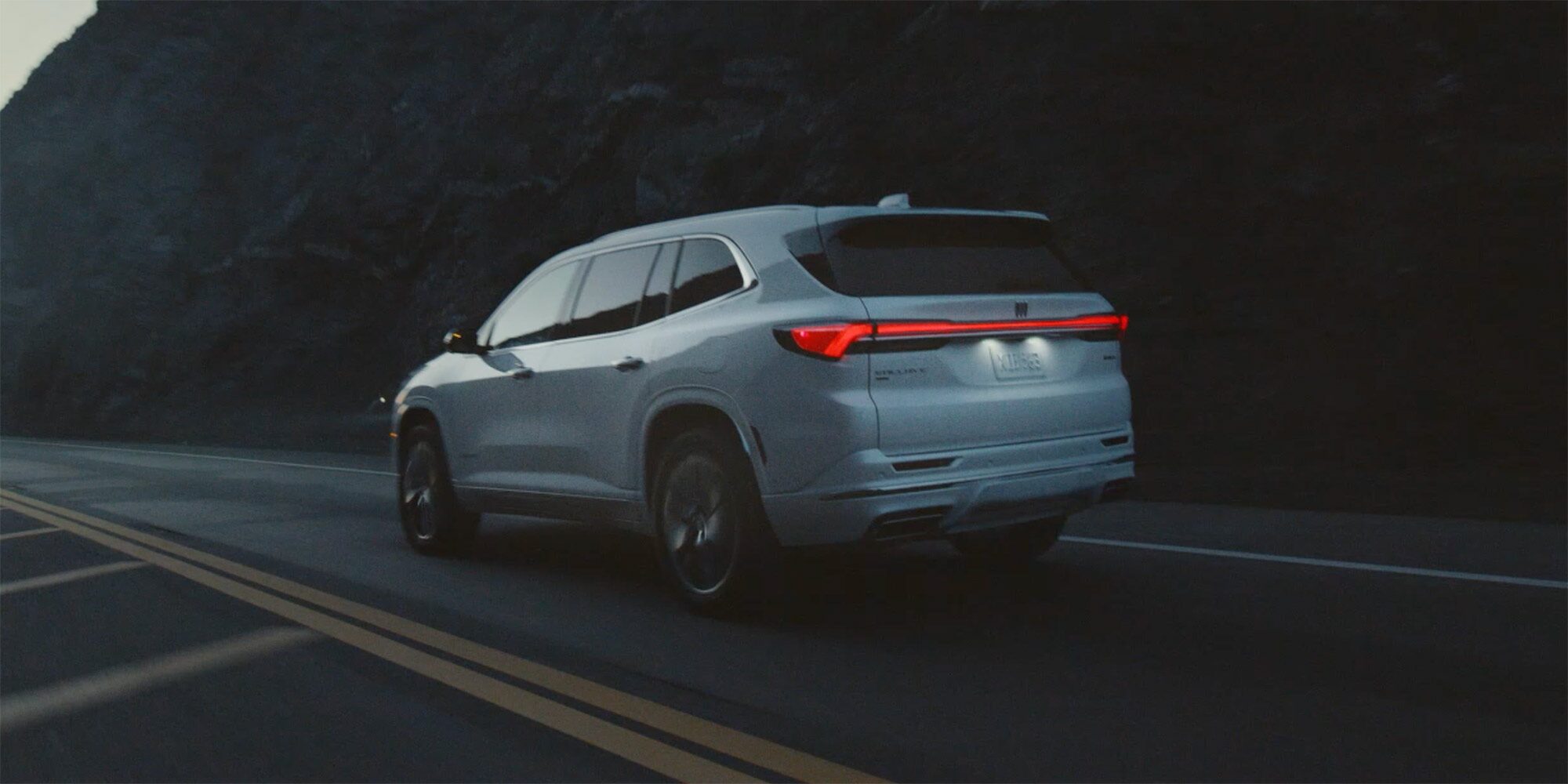 Three-Quarters Rear View of a 2026 White Buick Enclave Driving Down the Road Next to Mountains and a Body of Water