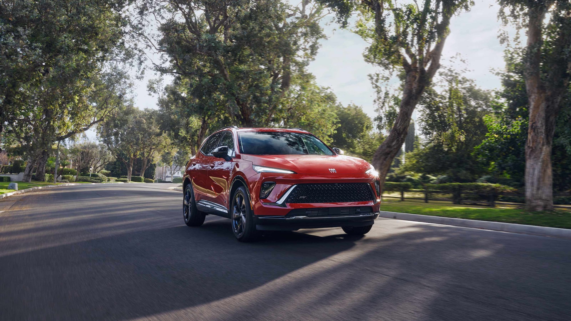 Front Three-Quarters View of the 2026 Buick Envision Compact SUV Driving a Road Lined with Green Trees